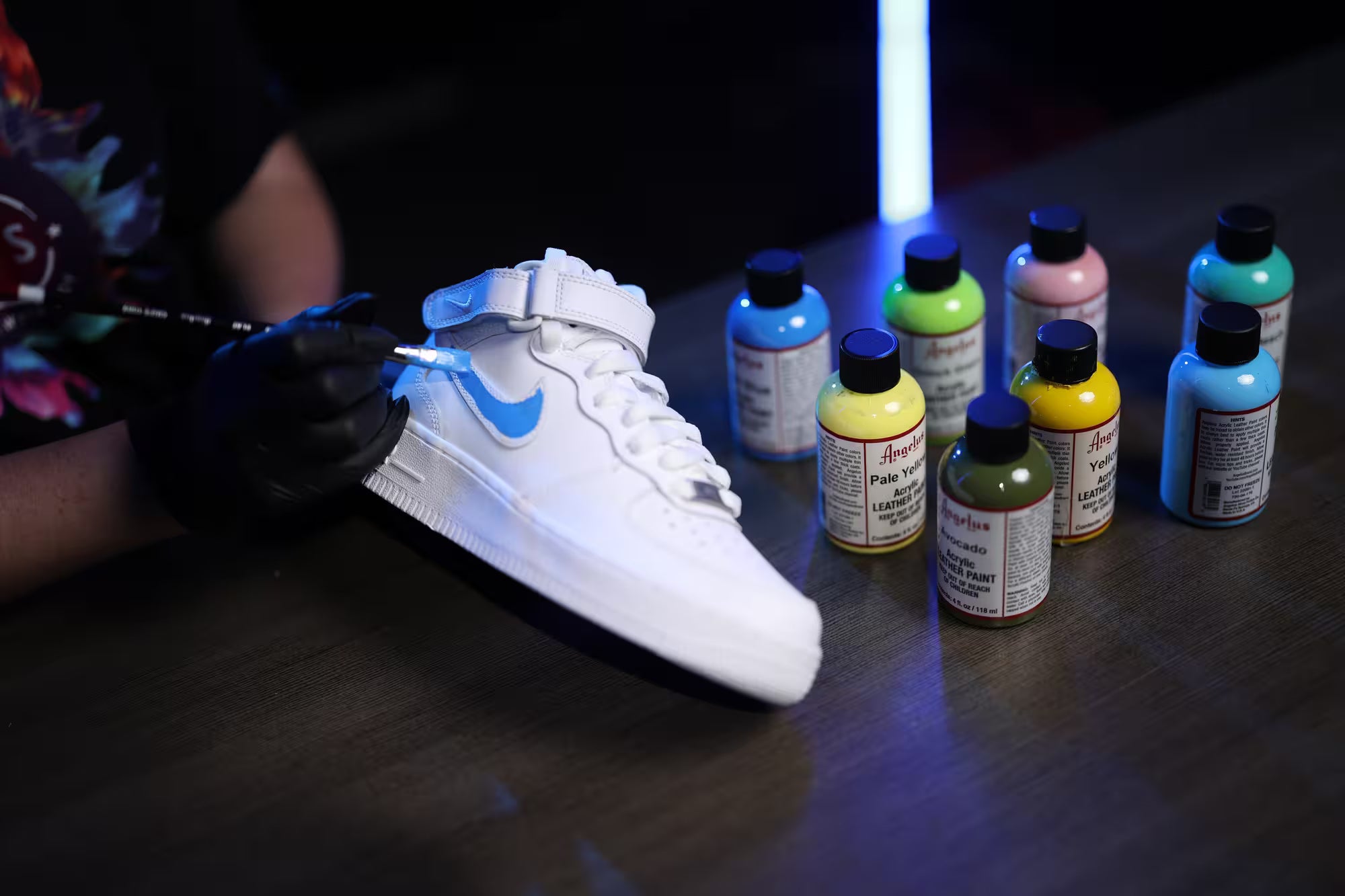 Joy working on a white sneaker with glowing blue design, surrounded by colorful bottles of paint.