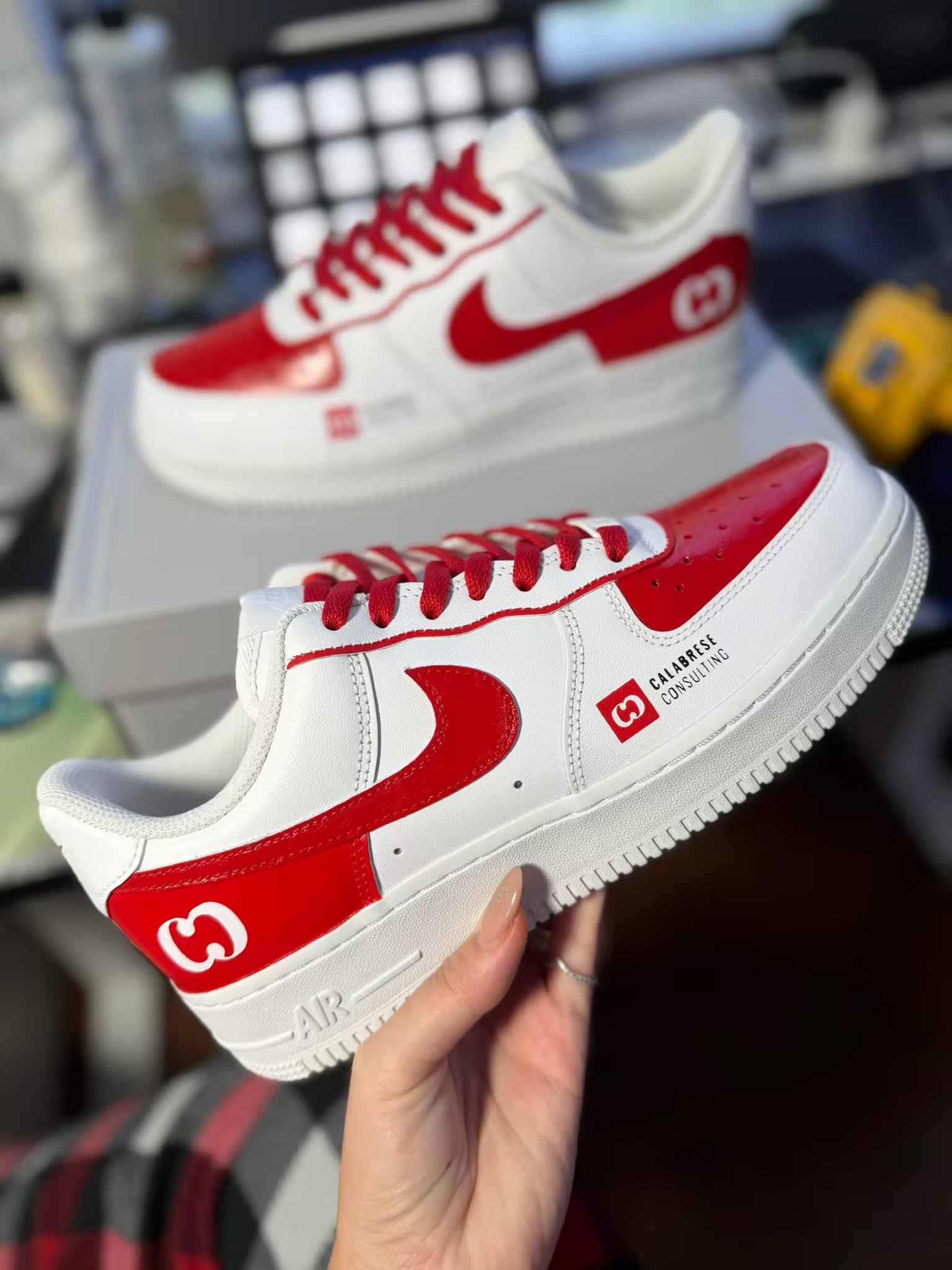 White and red sneakers with a brand logo held by a hand in an indoor setting.