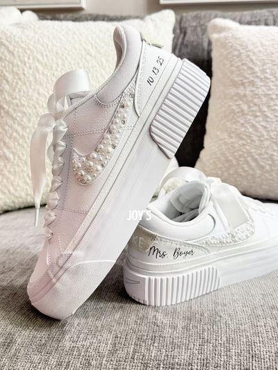 White custom wedding sneakers with pearl and rhinestone embellished swoosh, satin ribbon laces, and personalized bride name and wedding date on the heel