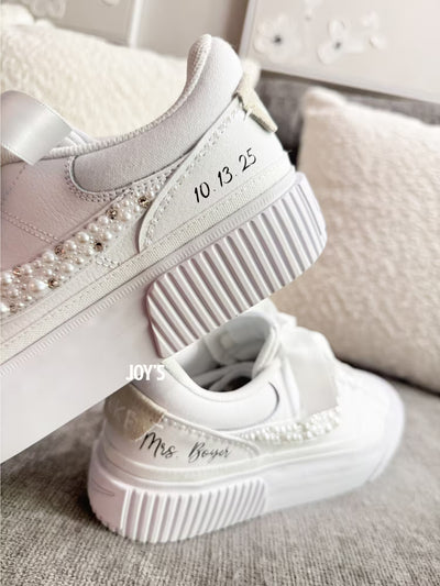 White custom wedding sneakers with pearl and rhinestone embellished swoosh, satin ribbon laces, and personalized bride name and wedding date on the heel