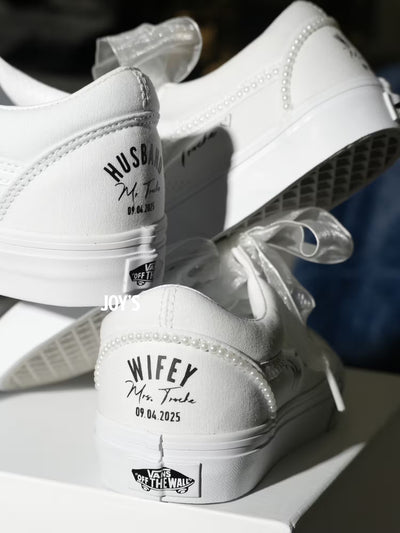 White custom wedding sneakers with pearl trim and personalized bride and groom names, wedding date, and “Wifey” and “Husband” heel text shown in a close-up detail view