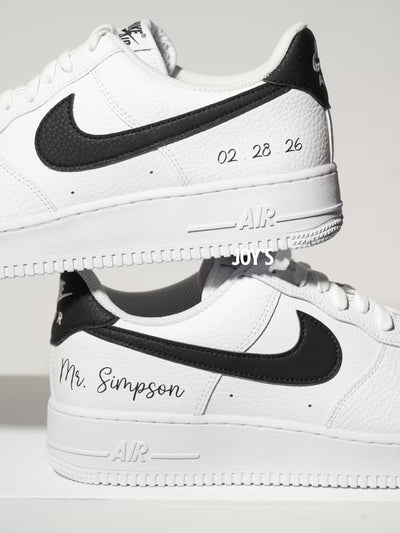 White custom Nike Air Force 1 sneakers with black swoosh, personalized last name and wedding date printed on the side, shown in a clean close-up view