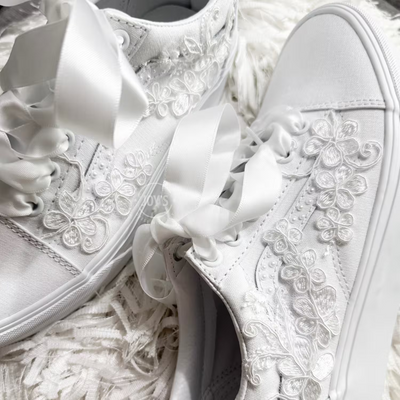 White platform Vans wedding sneakers with lace floral appliqués, pearl detailing, and satin ribbon laces, shown in a close-up side view