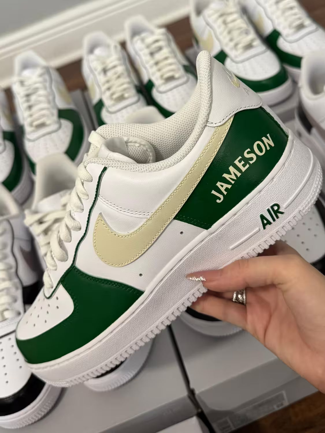 White Custom Jamson sneakers with green and yellow accents held by a hand, with more sneakers in the background.