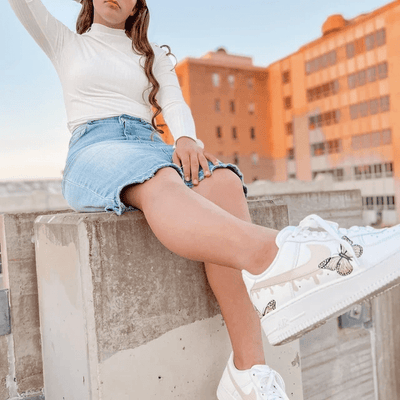 Lifestyle shot of a model wearing white custom Nike Air Force 1 sneakers with butterfly artwork, seated on a concrete ledge in an urban setting at sunset