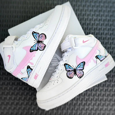 White Nike Air Force 1 Mid sneakers (standard sole, not platform), customized with hand-painted pastel butterflies in pink, blue, and lavender tones. Features soft pink swooshes with subtle drip accents, classic mid-top ankle strap, and a clean white base, photographed laid flat on a textured surface.