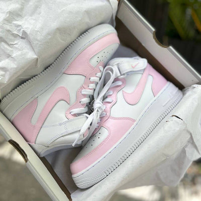 Pink and white **Nike Air Force 1 Mid** sneakers (non-platform), featuring soft pastel panels, classic ankle strap, and a clean mid-top silhouette, displayed outdoors on a white pedestal.
