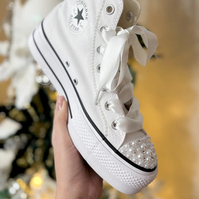 White platform Converse Chuck Taylor wedding sneakers with pearl-embellished toe cap and satin ribbon laces, shown in a hand-held close-up view