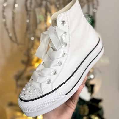 White platform Converse Chuck Taylor wedding sneakers with pearl-embellished toe cap and satin ribbon laces, shown in a hand-held close-up view