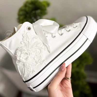 White platform Converse Chuck Taylor wedding sneakers with lace floral appliqués, satin ribbon laces, and personalized initials printed on the side, shown in a clean studio side view
