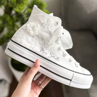 White platform Converse Chuck Taylor wedding sneakers with lace floral appliqués, satin ribbon laces, and personalized initials printed on the side, shown in a clean studio side view
