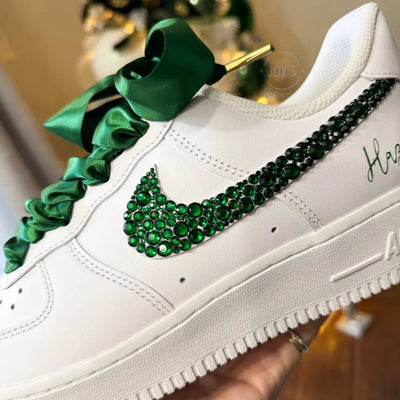 White custom Nike Air Force 1 sneakers with emerald green rhinestone swoosh, satin ribbon laces, and personalized name printed on the side, shown in a hand-held side view