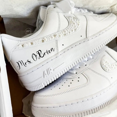Custom wedding white Nike Air Force 1 with pearl and rhinestone swoosh, white ribbon laces, and the name Mrs. personalized on the heel, shown held in hand with soft holiday lights in the background.