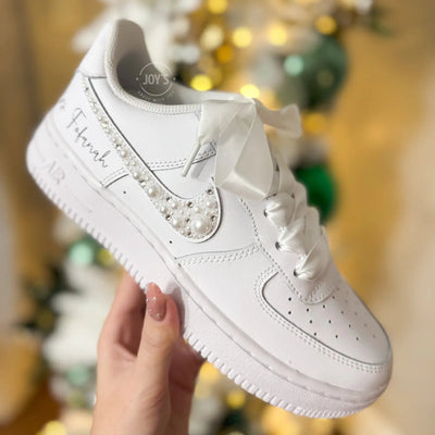 Custom wedding white Nike Air Force 1 with pearl and rhinestone swoosh, white ribbon laces, and the name Mrs. personalized on the heel, shown held in hand with soft holiday lights in the background.