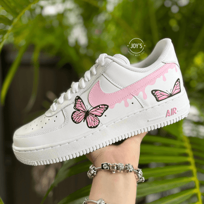 Custom white Nike Air Force 1 with pink drip swoosh and hand-painted pink butterfly details, shown held in hand against a green foliage background.