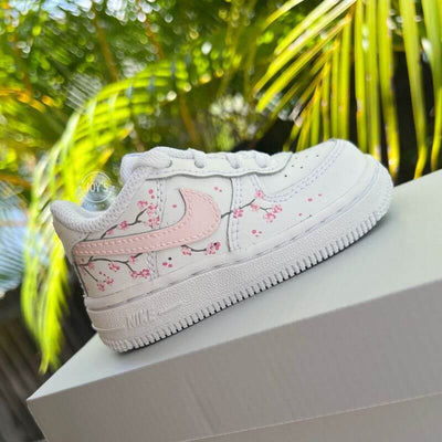 White Nike Air Force 1 toddler sneakers with soft pink swoosh and cherry blossom branch details, shown from the side.