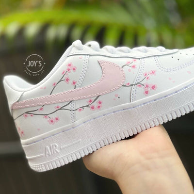 White Nike Air Force 1 with soft pink swoosh and hand-painted cherry blossom branches, shown in close-up.