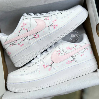 White Nike Air Force 1 with soft pink swoosh and hand-painted cherry blossom branches, shown in close-up.