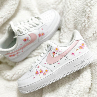 White Nike Air Force 1 sneaker with hand-painted pink cherry blossom flowers and a soft pink swoosh, shown close-up on a plush white background.
