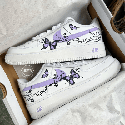 Pair of custom white Nike Air Force 1 sneakers in a shoebox, featuring lavender swooshes with black floral vines and hand-painted purple butterfly designs.