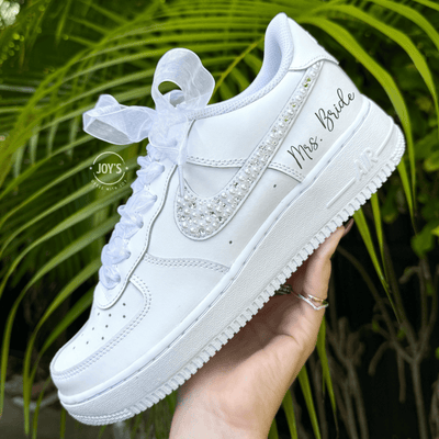 Custom wedding white Nike Air Force 1 with pearl and rhinestone swoosh, white ribbon laces, and the name Mrs. personalized on the heel, shown held in hand with soft holiday lights in the background.