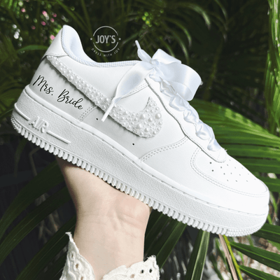 Custom white wedding Nike Air Force 1 with pearl-embellished swoosh, white ribbon laces, and "Mrs. Bride” on the side shown held in hand